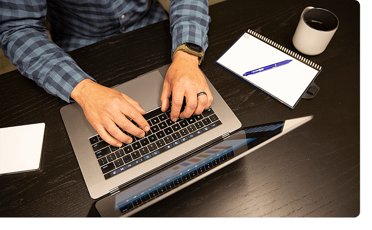 Man working on a laptop with a notepad and coffee cup beside him.